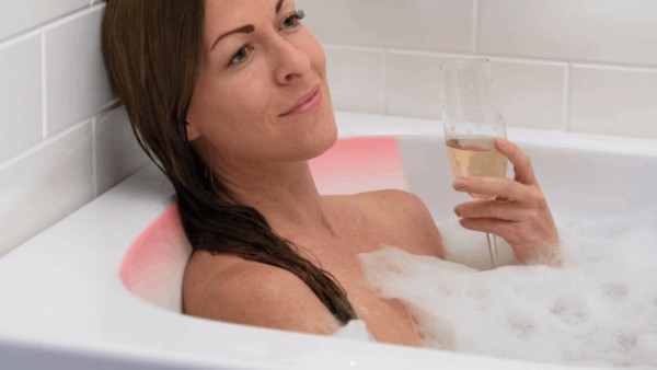 Woman enjoying bubble bath leaning on illuminated heated backrest while sipping champagne